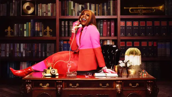 Big Freedia, in a vibrant pink and orange suit, sits at a desk, talking on a vintage phone. Her success is evident in the opulent office, hinting at her impressive big Freedia net worth.
