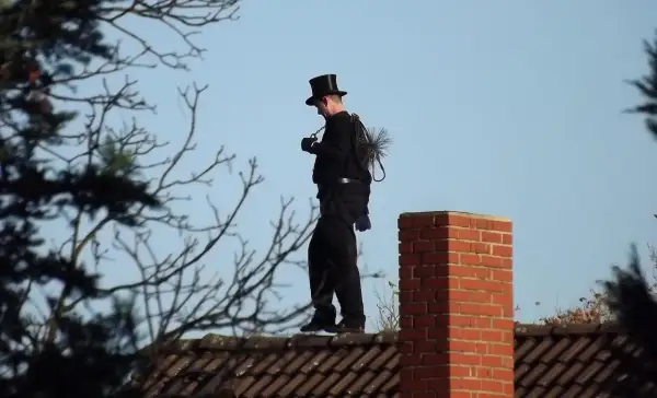 Ein Schornsteinfeger in Zylinder und schwarzer Kleidung steht auf einem Dach. Er trägt seine Bürste und Kette bei sich. Man fragt sich, wie oft kommt der Schornsteinfeger wohl?