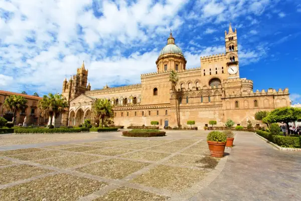 La maestosa cattedrale di Palermo si erge sotto un cielo azzurro con nuvole bianche, con la sua architettura imponente e il suo cortile lastricato.