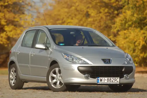 Srebrny Peugeot 307 z widocznymi żarówkami reflektorów, zaparkowany na tle jesiennych drzew.
