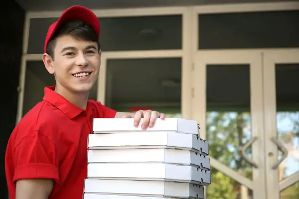 Uśmiechnięty pracownik młodociany w czerwonej koszulce i czapce niesie stos pudełek z pizzą.