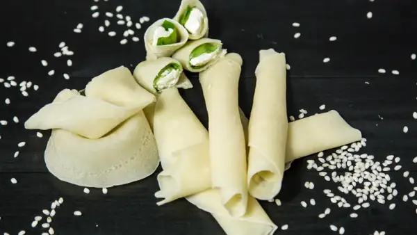 Rotolini di crepes con farina di riso, ripieni di formaggio fresco e basilico, sparsi su un tavolo scuro con semi di sesamo.