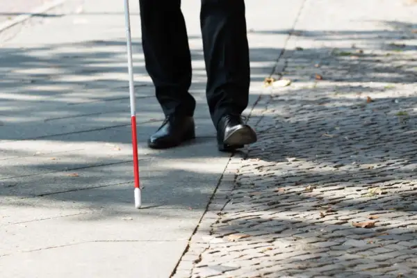 Un uomo con bastone bianco e rosso cammina su un marciapiede, un valido accompagnamento per la cecità parziale.