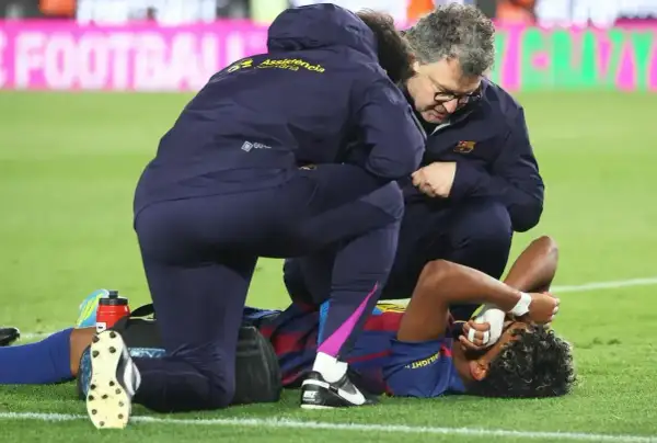 Piłkarz leży na boisku, trzymając się za twarz. Personel medyczny udziela mu pomocy, obawiając się poważnej kontuzji.