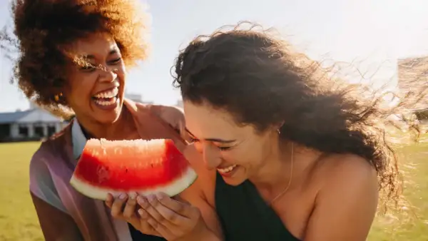 Zwei lachende Frauen teilen ein Stück Wassermelone. Perfekt für einen heißen Tag, da Wassermelone kalorienarm ist und erfrischt.