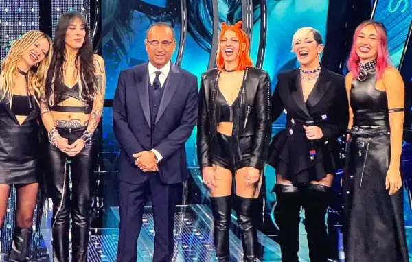 Micky Paiano con un completo scuro, affiancato da quattro donne con outfit in pelle audaci e colorati, sorridono sul palco.