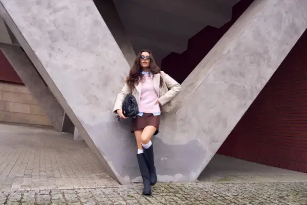 Dziewczyna w różowym sweterku do brązowej spódniczki, beżowej kurtce i czarnych kozakach pozuje na tle betonowej konstrukcji.