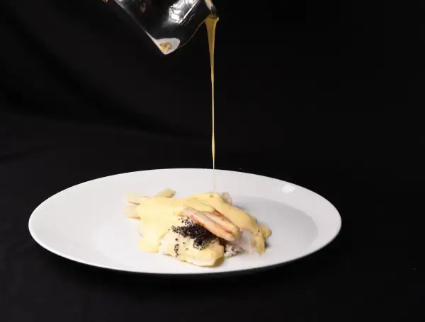 Ein Koch übergießt Fisch mit Spargel und Kaviar mit einer cremigen Sauce. Die Sauce sieht aus, als wäre die Sahne geronnen, was dem Gericht eine interessante Textur verleiht.