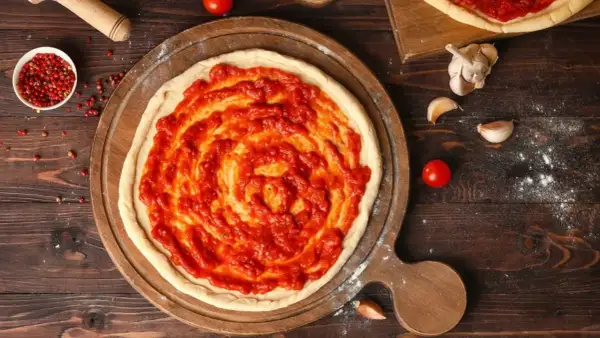 Une pâte à pizza crue est étalée sur une planche en bois, recouverte d'une sauce tomate maison. La question quelle sauce tomate pour pizza est au cœur de cette préparation.
