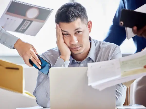 Zmęczony mężczyzna przy laptopie, otoczony dokumentami i telefonami. Czy to właśnie multitasking co to znaczy?