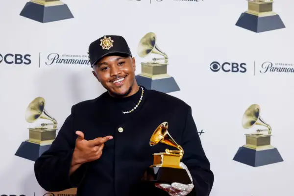 Khris Riddick-Tynes smiles, holding a Grammy award and pointing to it. He wears a black cap and jacket.