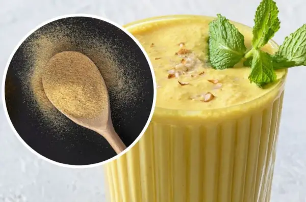 Puszysty mango lassi w szklance, ozdobiony miętą i orzechami. Obok łyżka z proszkiem, być może przyprawą do mango lassi.