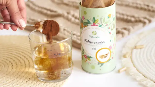 Łyżeczka z proszkiem ashwagandhy wsypywana do szklanki wody. Opakowanie Naturalis Ashwagandha BIO obok.