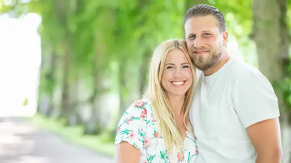 Ett leende par, Nanna Olasdotter Hallberg och hennes partner, njuter av en sommardag utomhus bland gröna träd.