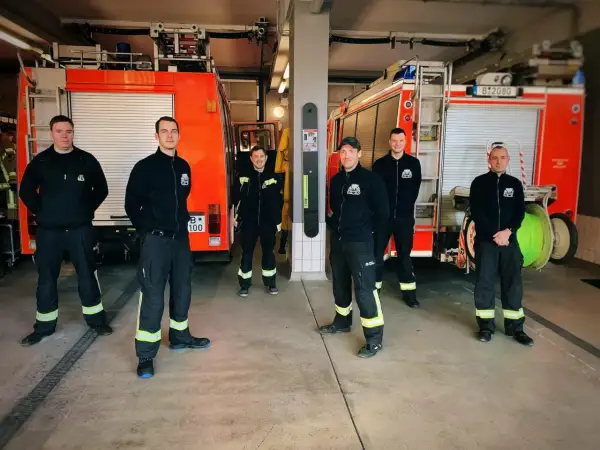 Sechs Männer in Einsatzkleidung stehen vor zwei Feuerwehrautos. Sie leisten ehrenamtlich Dienst bei der Feuerwehr und sind bereit für den nächsten Einsatz.