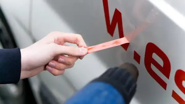 Hand zieht roten Aufkleber vom Auto ab, während ein Heißluftföhn das Entfernen erleichtert.
