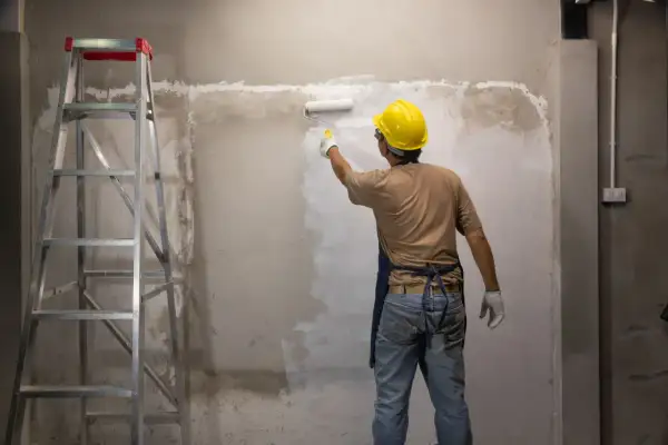 Operaio con casco giallo stende le vernici bianche su un muro grezzo, accanto a una scala metallica.