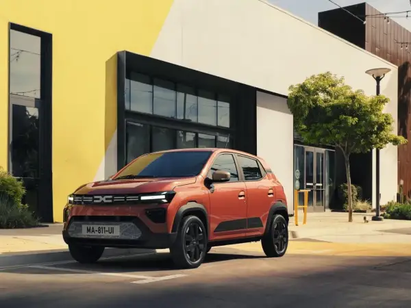 Pomarańczowy Dacia Spring Electric zaparkowany przed nowoczesnym budynkiem z żółtymi akcentami.