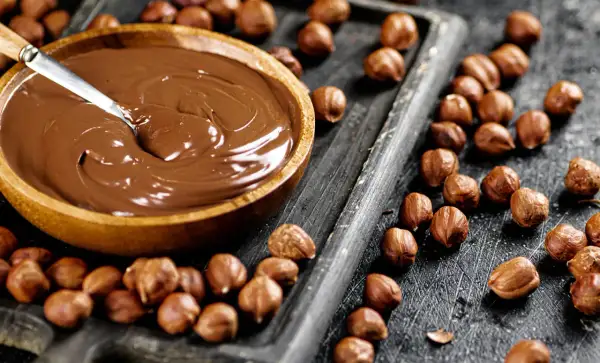 Schokocreme selber machen: Eine Schüssel voller cremiger Schokocreme neben gerösteten Haselnüssen auf einem Holzbrett.