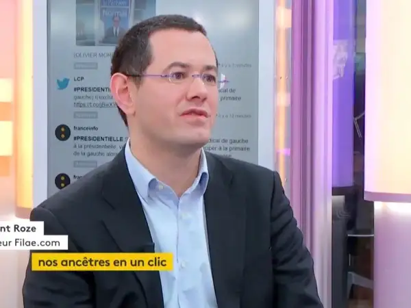 Un homme en costume, lunettes, parle. Le nom de famille gratuit "Filae.com" est visible, suggérant une recherche d'ancêtres.