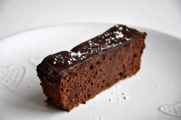 Wilgotny brownie z fasoli, polany czekoladą i posypany cukrem pudrem, na białym talerzu z serduszkami.
