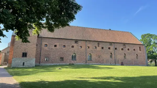 Imponerande tegelbyggnad med tegeltak, en del av Vadstena kloster, omgiven av grönskande gräsmatta och träd under en klarblå himmel.