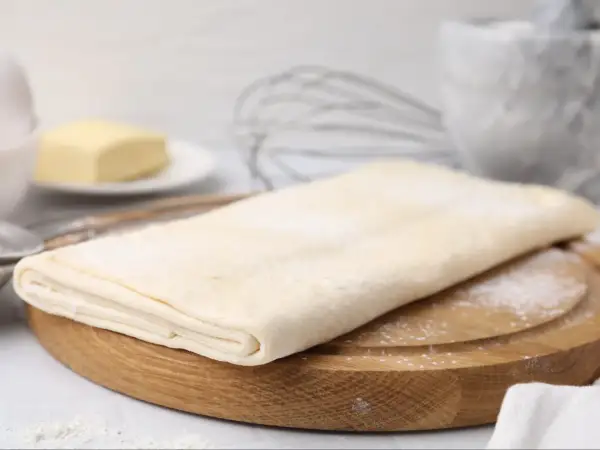 Gotowe ciasto francuskie na desce, obok masło i trzepaczka. Sprawdź, jaka jest ciasto francuskie cena!