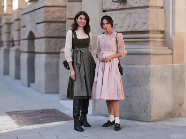 Zwei Frauen posieren vor einer Steinfassade. Eine trägt ein grünes Dirndl mit weißer Bluse und schwarze Stiefel.