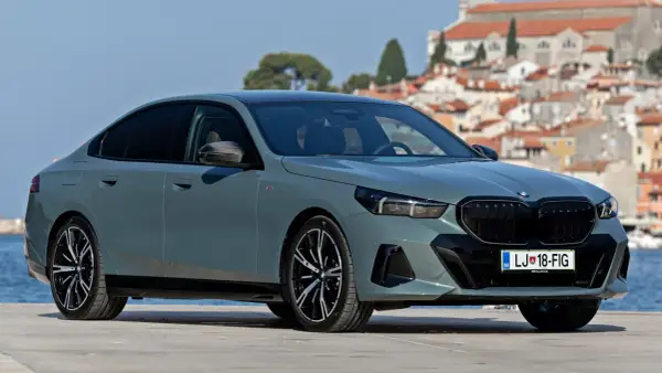 Szary sedan BMW 5 2025 z czarnym grillem i felgami, zaparkowany na tle malowniczego miasta nad morzem.