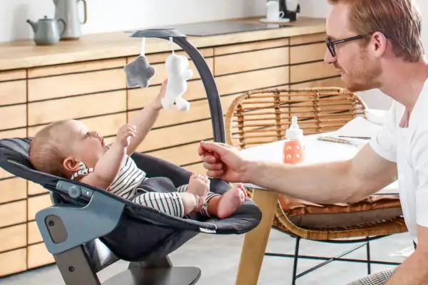 Ein Vater spielt mit seinem Baby in einem Hochstuhl. Ab wann Babys im Hochstuhl sitzen, hängt von ihrer Entwicklung ab.