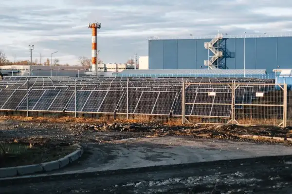 Duże pole paneli słonecznych przed niebieskim budynkiem. W tle pomarańczowo-biała wieża. THD.