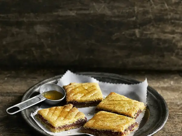 Quatre makrouts algériens sur une assiette, arrosés de miel. Une cuillère doseuse remplie de miel est à côté. Parfait pour une recette makrout algérien facile.