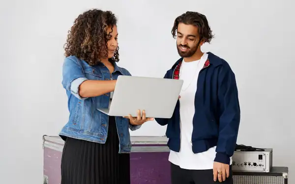 A woman and man review a laptop, discussing music artist management. They stand near stage equipment.