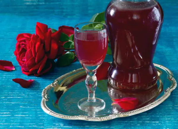 Aromatic nalewka z płatków róży w kieliszku i karafce, obok czerwona róża i płatki na srebrnej tacy.