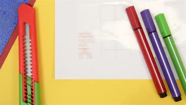 Un cutter rouge et trois feutres (rouge, violet, vert) sont prêts pour un projet créatif. Une **lettre noire** est visible sur le papier de fond.