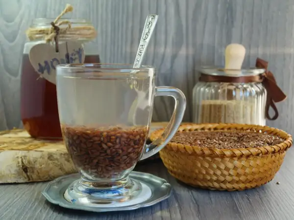 Szklanka z wodą i siemieniem lnianym, obok miód i koszyk z nasionami. Kiedy pić siemię lniane rano czy wieczorem? To pytanie o zdrowie.