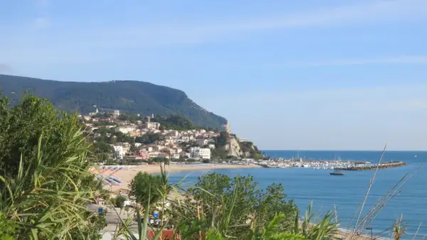 Vista panoramica della Riviera del Conero con un borgo arroccato su una collina, spiaggia attrezzata e un porto turistico.
