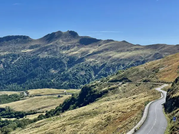 Route sinueuse dans les montagnes, avec des pâturages dorés et une forêt dense. Pas de Peyrol, un paysage grandiose sous un ciel bleu.