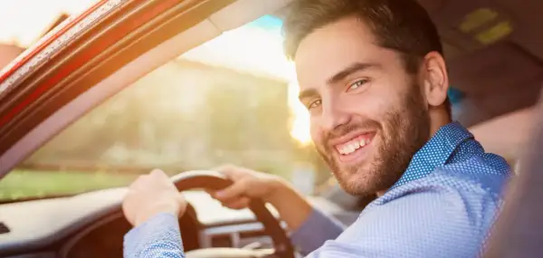 Un homme souriant au volant de sa voiture, prêt pour sa previsite contrôle technique. Le soleil brille, annonçant une journée sereine.