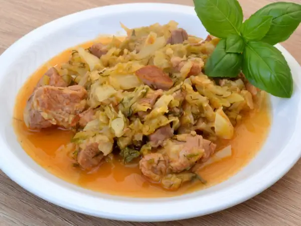 Pyszny bigos z młodej kapusty z kawałkami mięsa, podany na białym talerzu, ozdobiony gałązką bazylii.