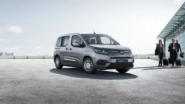 Srebrna toyota 7 osobowa czeka na pasażerów przy lotnisku. Ludzie z walizkami idą w stronę samochodu.