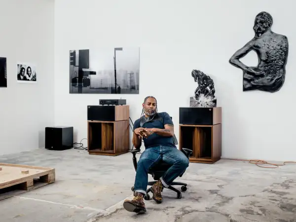 A man sits in an office chair in a gallery space, surrounded by art and audio equipment, a scene that feels like a snapshot of Arthur Jafa's creative process.