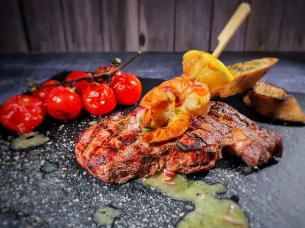 Perfektes Surf and Turf Rezept: saftiges Steak mit gegrillten Garnelen, Kirschtomaten und Zitronenspalte auf schwarzem Stein.