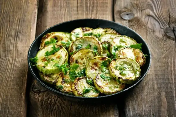 Zucchine marinate cotte, grigliate e guarnite con prezzemolo fresco, servite in una padella su un tavolo di legno rustico.