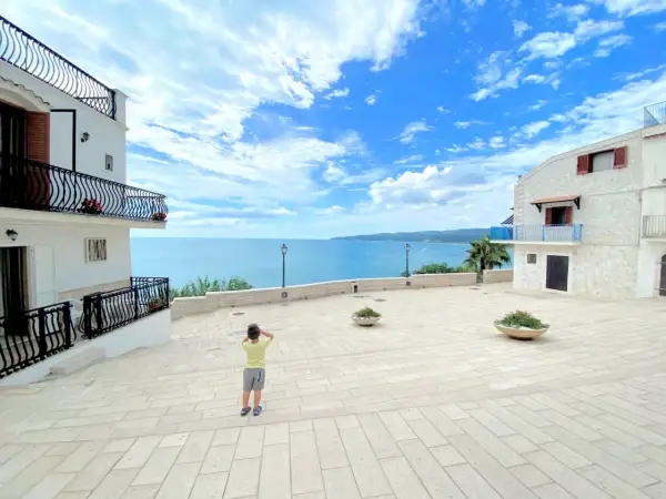 Bambino ammira il mare azzurro dalla piazzetta Petrone a Vieste, con case bianche e cielo nuvoloso.