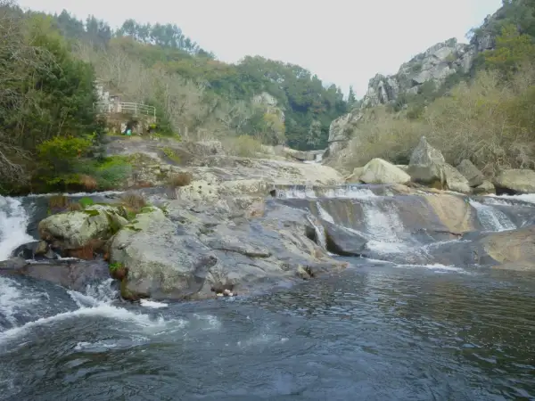 A fervenza de Segade, con auga caendo sobre rochas e un pequeno refuxio na ladeira verde.
