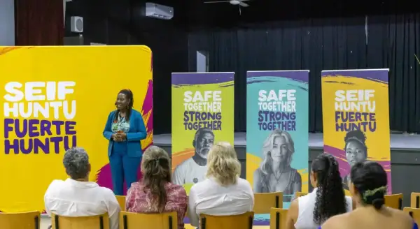 Een spreker staat voor een gele banner met "SEIF HUNTU FUERTE HUNTU". Achter haar staan banners met "SAFE TOGETHER STRONG TOGETHER". Dit is een evenement voor hss sw jeugd.
