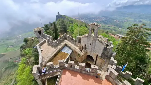 Widok z góry na kamienne fortyfikacje i wieże podczas san marino roaming. W oddali widać mgliste wzgórza i miasteczko.