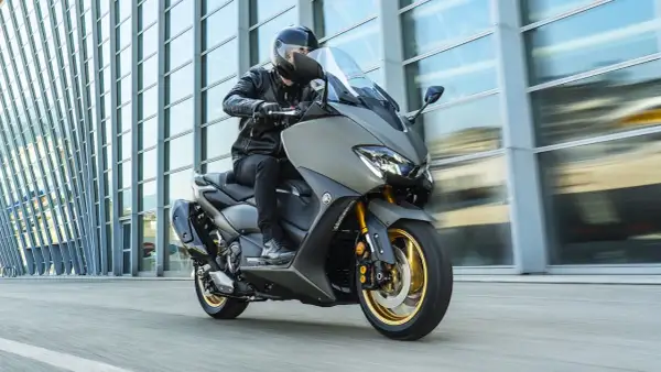 Hombre en moto clásica moderna, con chaqueta de cuero, circulando frente a un edificio de cristal.