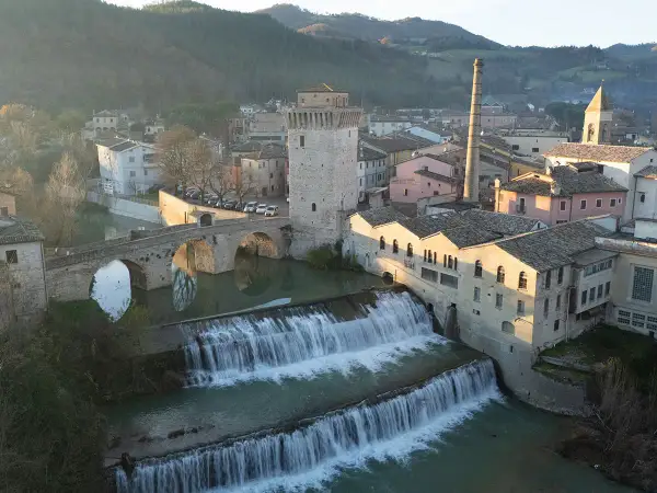 Cascata e ponte ad arco a Fermignano, vicino Urbino, con edifici storici e una ciminiera.